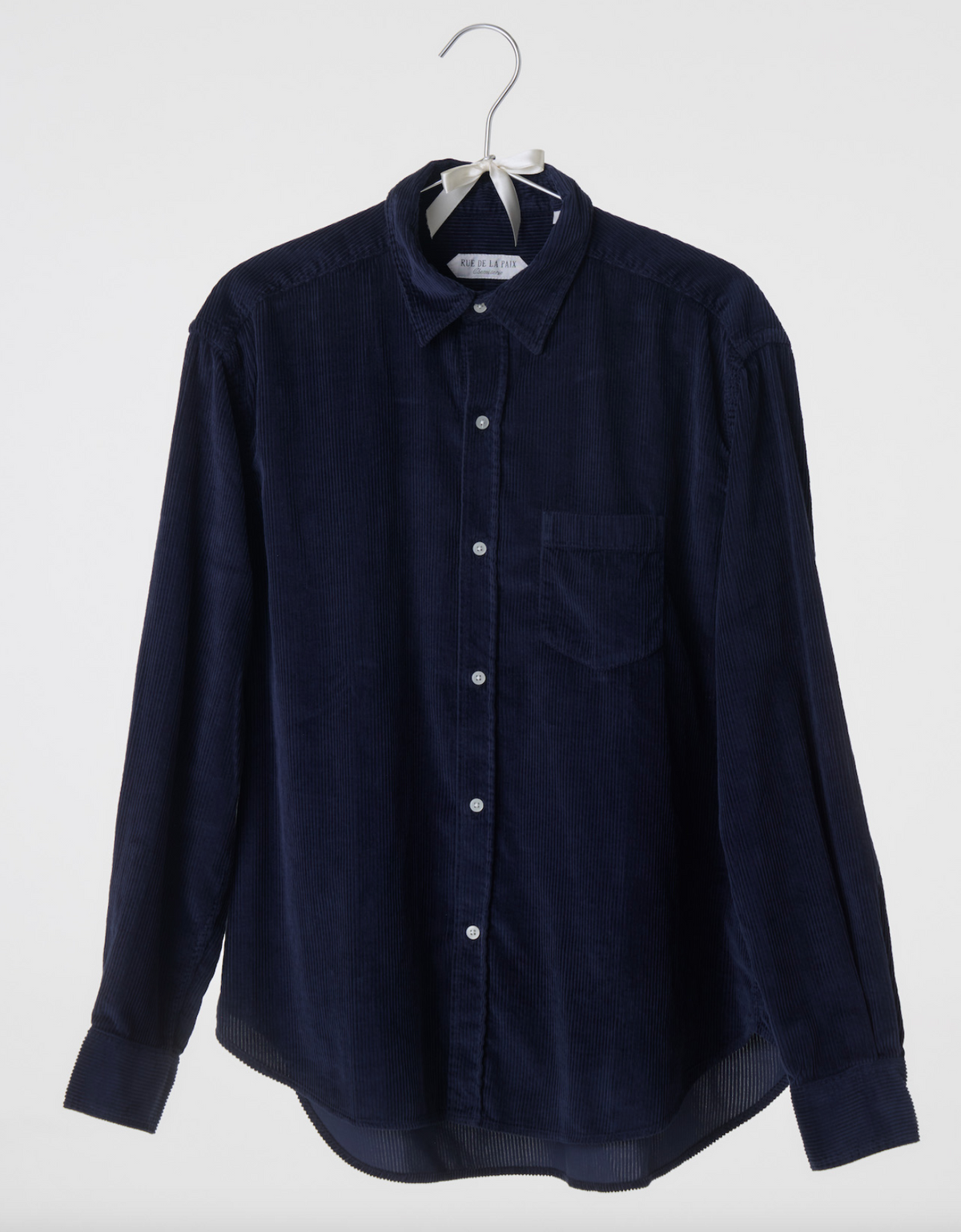 Rue de la Paix La Garçonne Shirt in navy corduroy on a hanger, with a classic collar, white buttons, chest pocket and curved hem